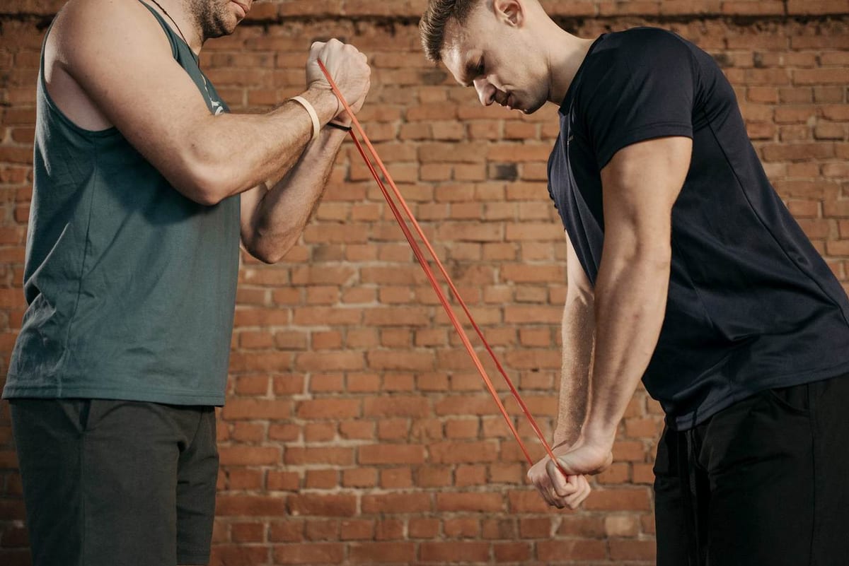 A man pulls a red resistance band held steady by his trainer in front of a brick wall