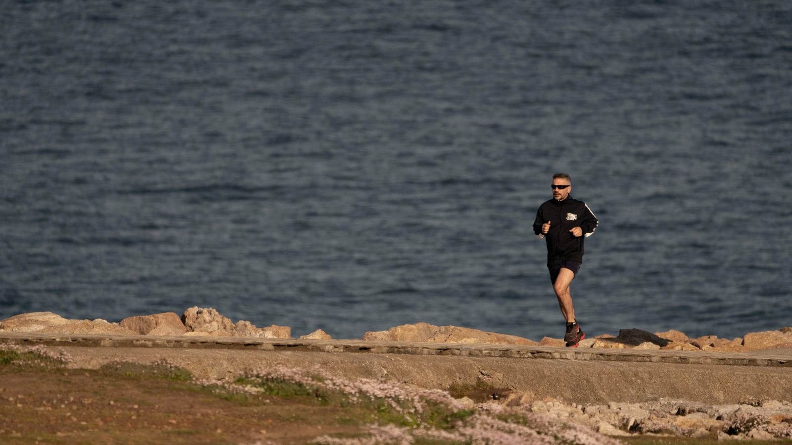A man runs along a rocky coastal path beside a calm blue sea