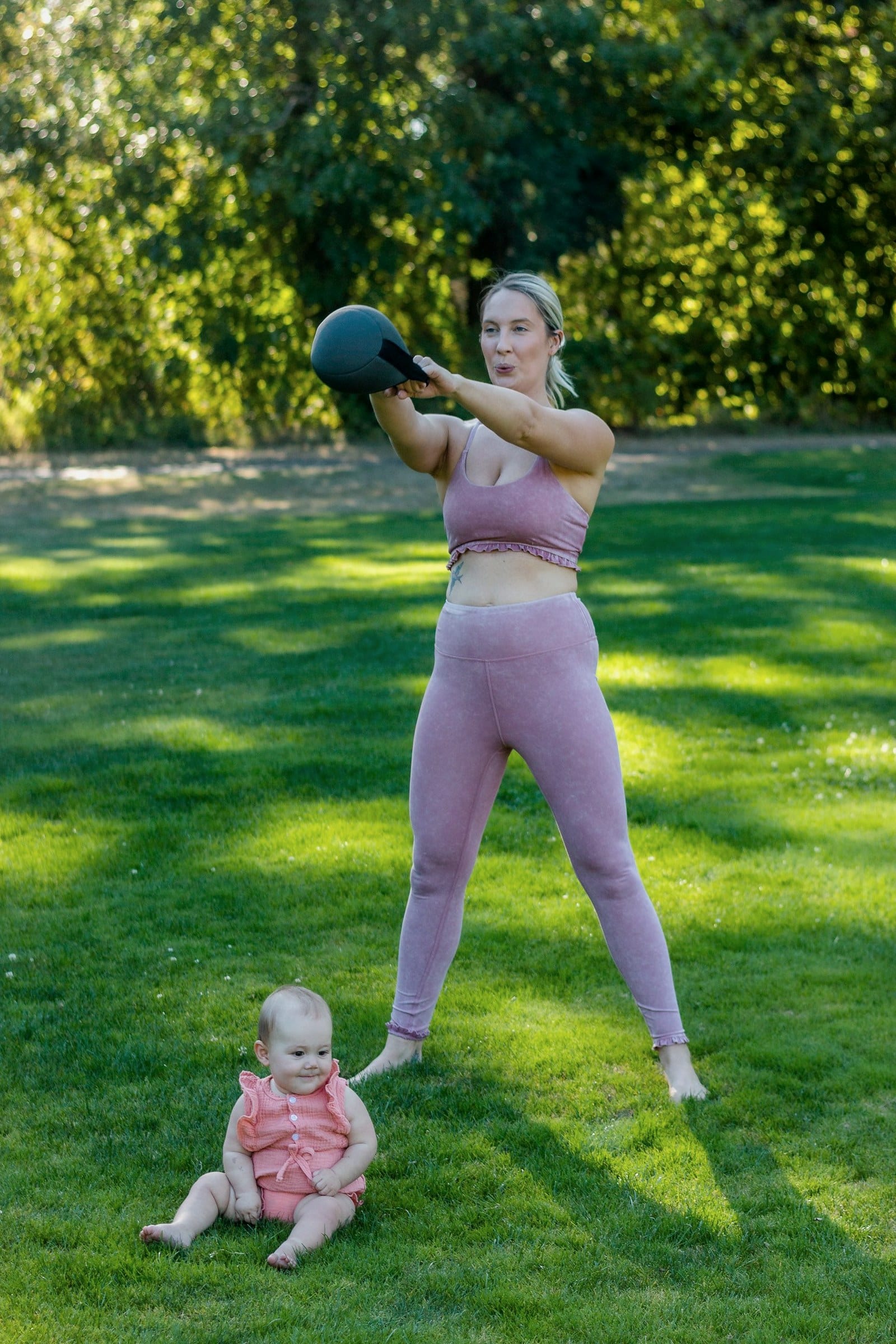 A mother swings a kettlebell on a grassy lawn while her baby sits nearby