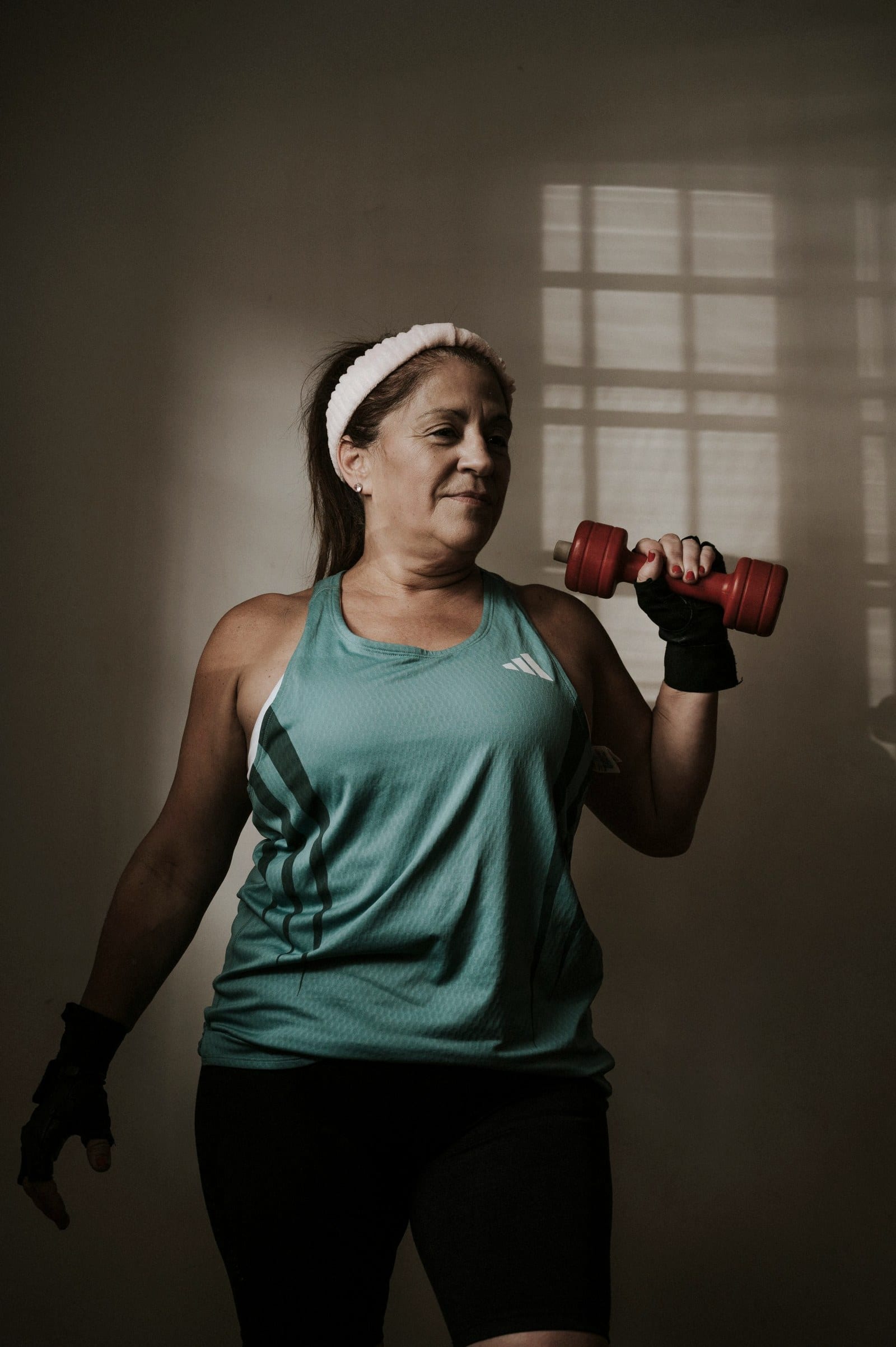 A woman in a teal top lifts a red dumbbell beside a sunlit wall