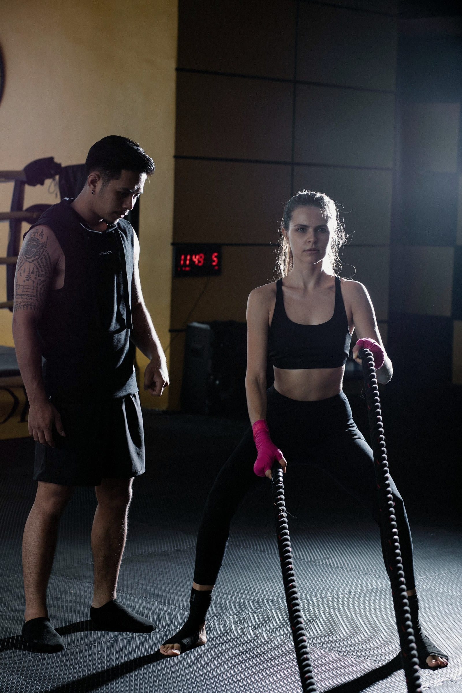 A woman with pink hand wraps holds battle ropes while her coach watches in a dark gym