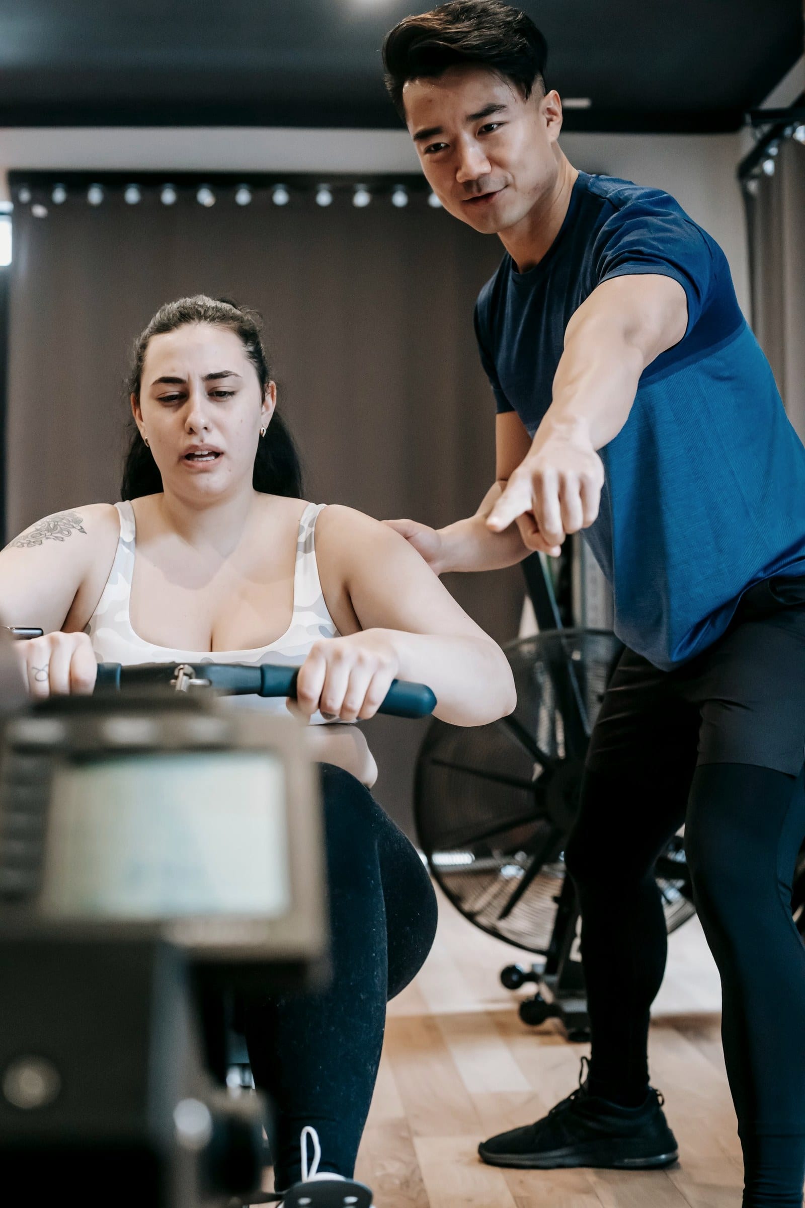 A trainer stands beside a woman rowing on a machine, guiding her effort in a gym