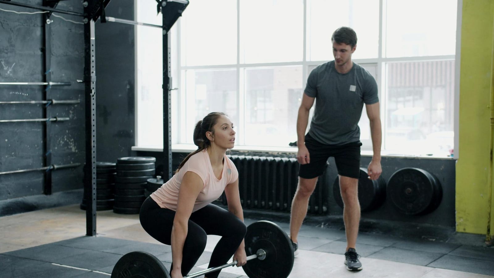 A woman performs deadlifts while her trainer watches in a bright gym