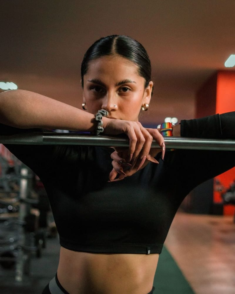 A woman personal trainer leans on a barbell in a dim gym, looking at the camera