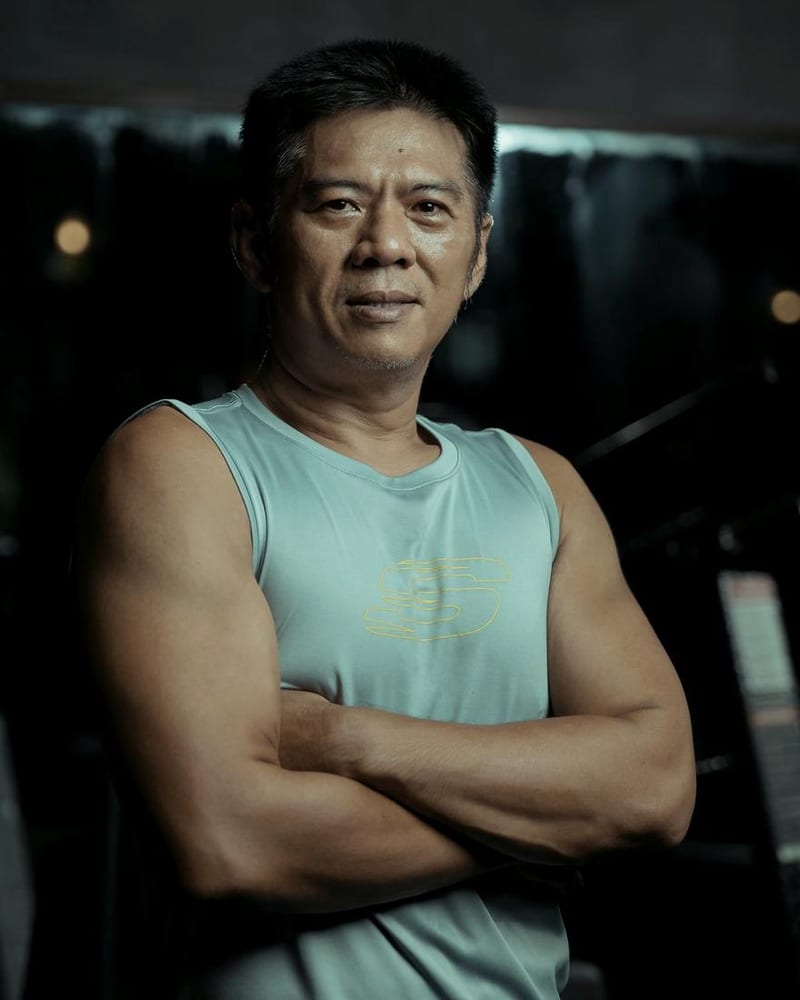 A mature man in a light blue vest stands with crossed arms in a gym