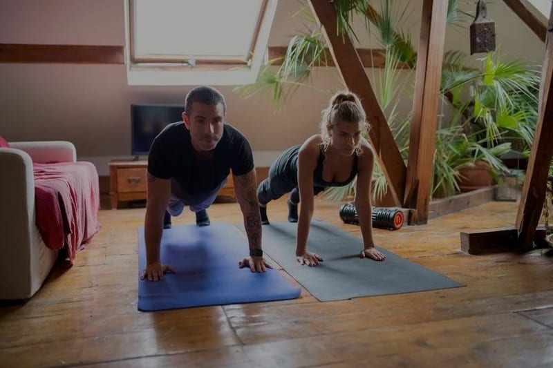 A man and woman do push-ups side by side on yoga mats in a cosy home attic room