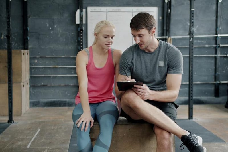 A personal trainer writes on a clipboard while discussing a plan with a seated client in a gym