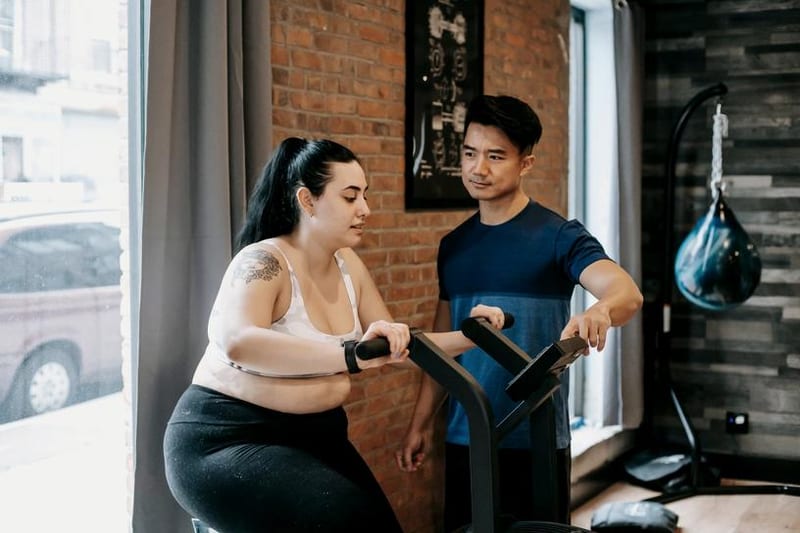 A woman pedals an exercise bike while her trainer adjusts the settings in a gym with brick walls