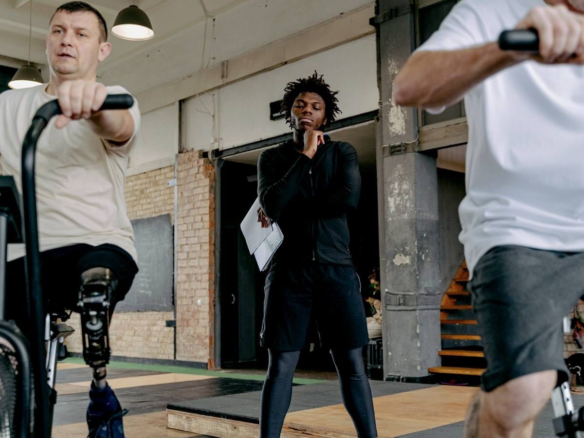A trainer holding a clipboard oversees two clients cycling in a gym