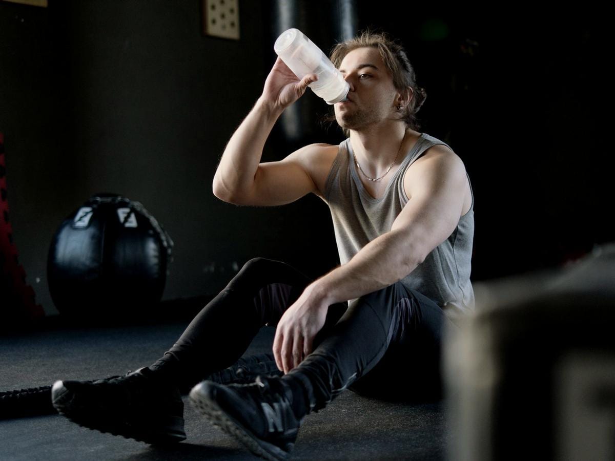 A man in gym kit sits on the floor drinking from a water bottle between sessions