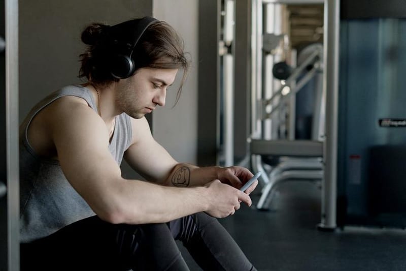 A man wearing headphones checks his phone while sitting in a gym