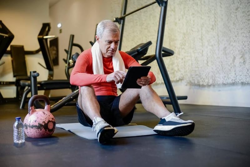 An older man in a red top reviews a tablet while sitting on a gym mat